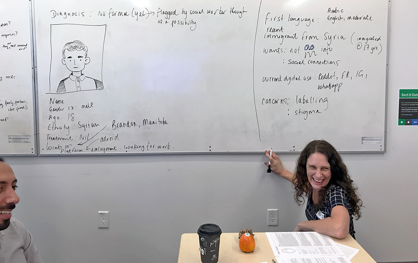 Erin does some whiteboarding while Aidin smiles. She's filling out a persona template for an 18-year old Syrian immigrant.