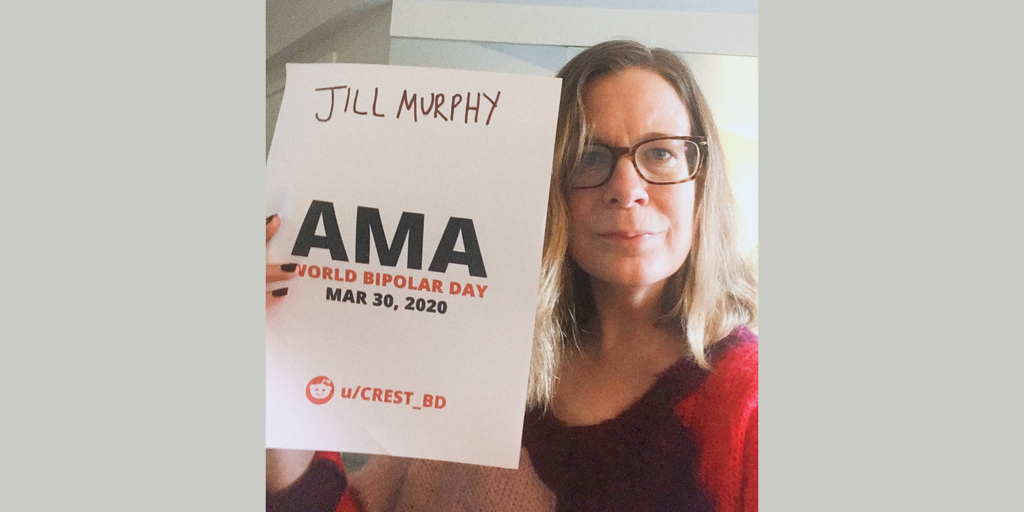 A close-up of Jill in her office, wearing a red sweater and holding the proof sign.