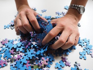 A white person's hands scooping up a pile of blue and purple puzzle pieces that are scattered on a white table. The person is wearing a watch.