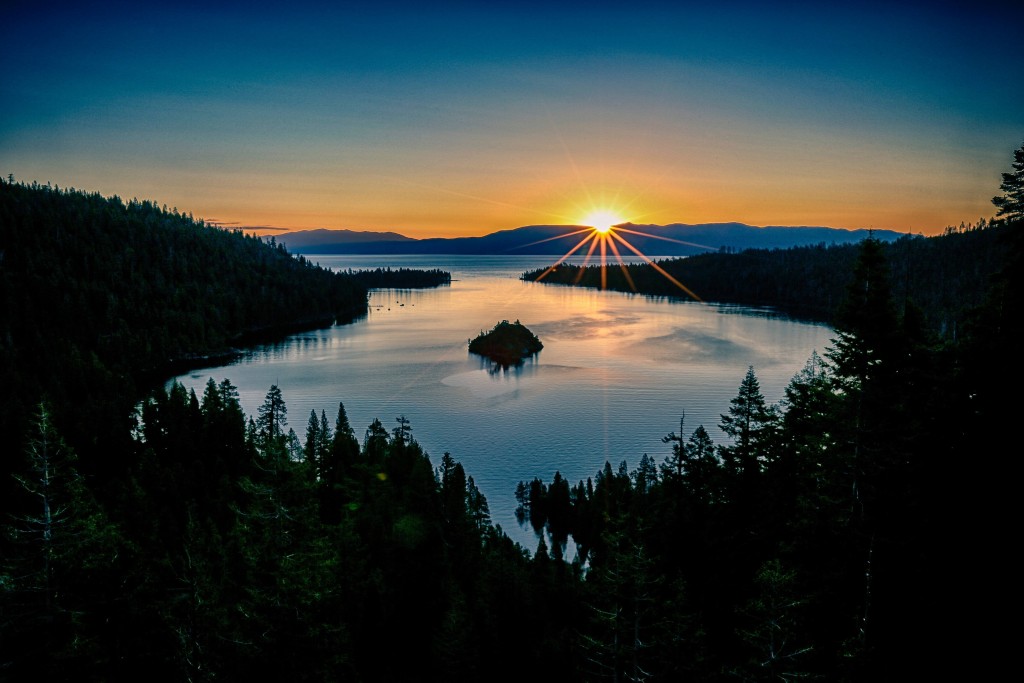 A burst of sunlight peeks out from behind low mountains. The light of dawn extends across a wide and clear lake. In the foreground, evergreen trees are silhouetted against the brightness of the lake.