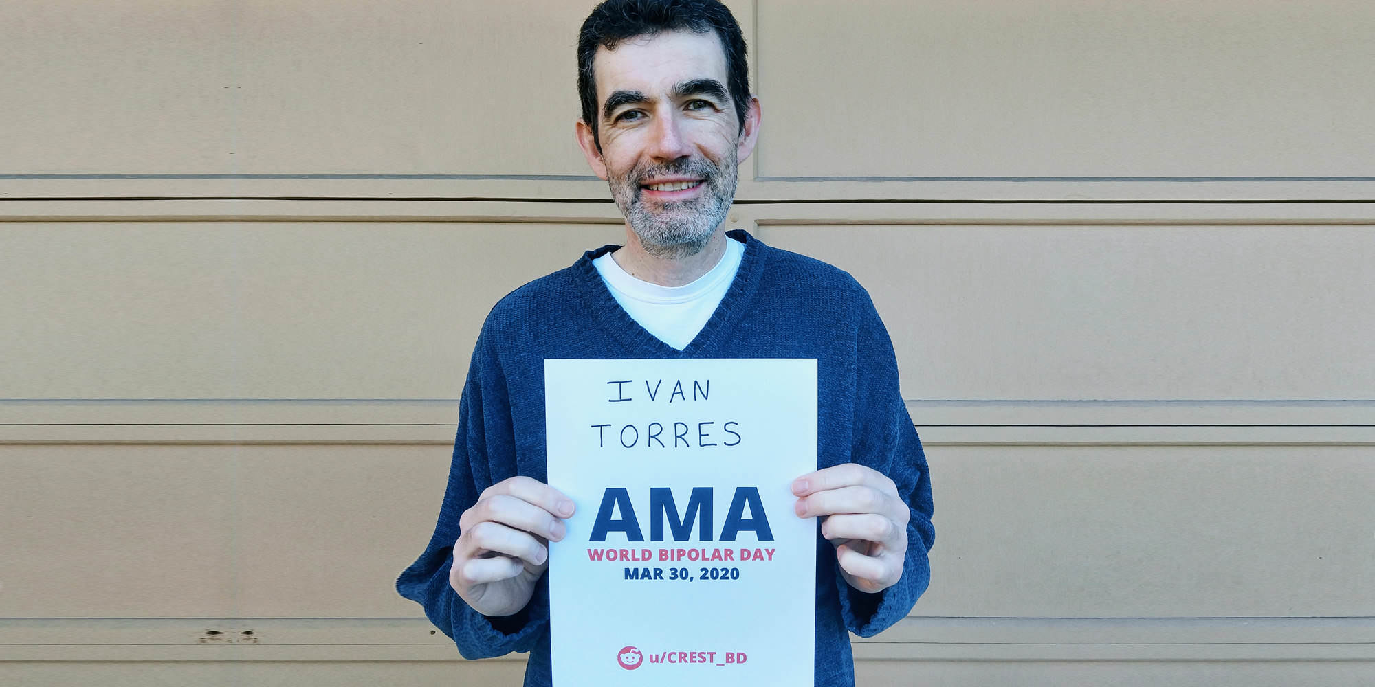 Ivan wearing a blue sweater, standing in front of a garage door and holding his proof side.