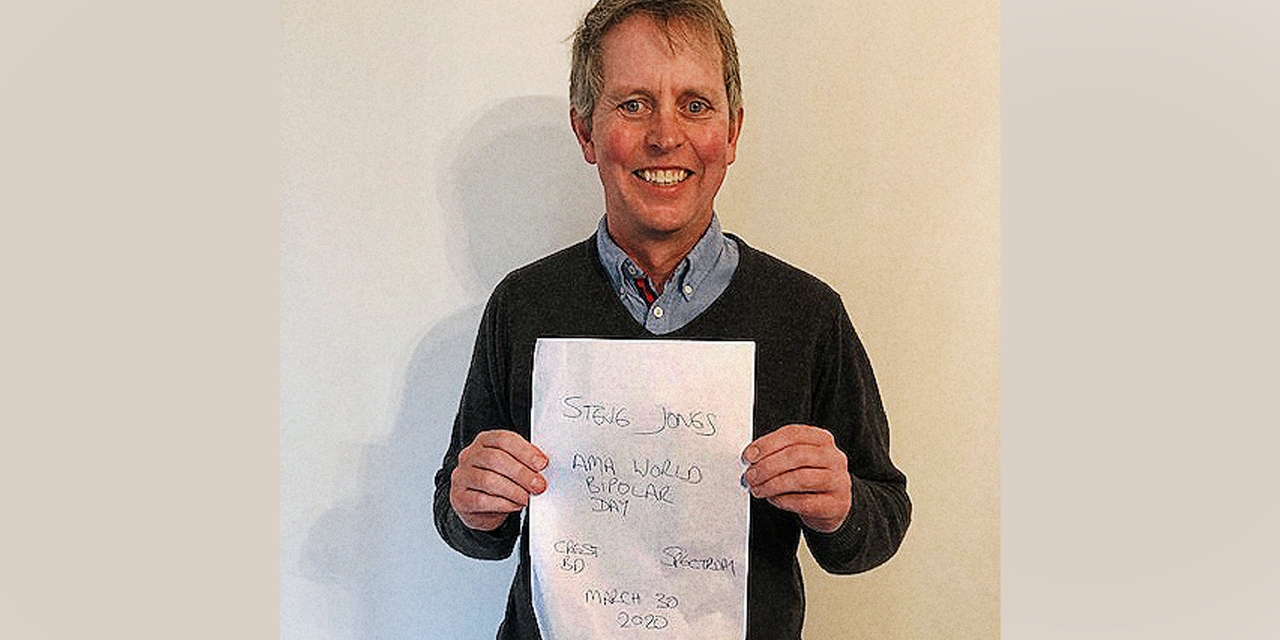 Steven Jones, smiling and wearing a darkly-coloured sweater standing against a beige wall and holding a proof sign.