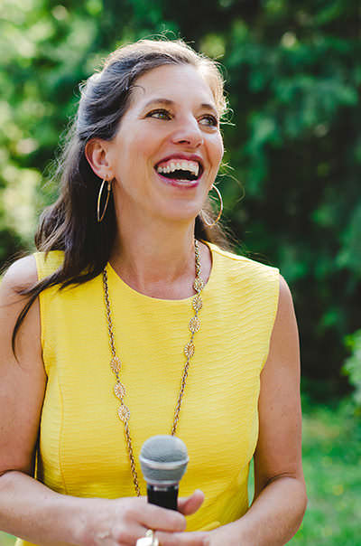 Victoria dressed in yellow, holding a microphone and laughing.