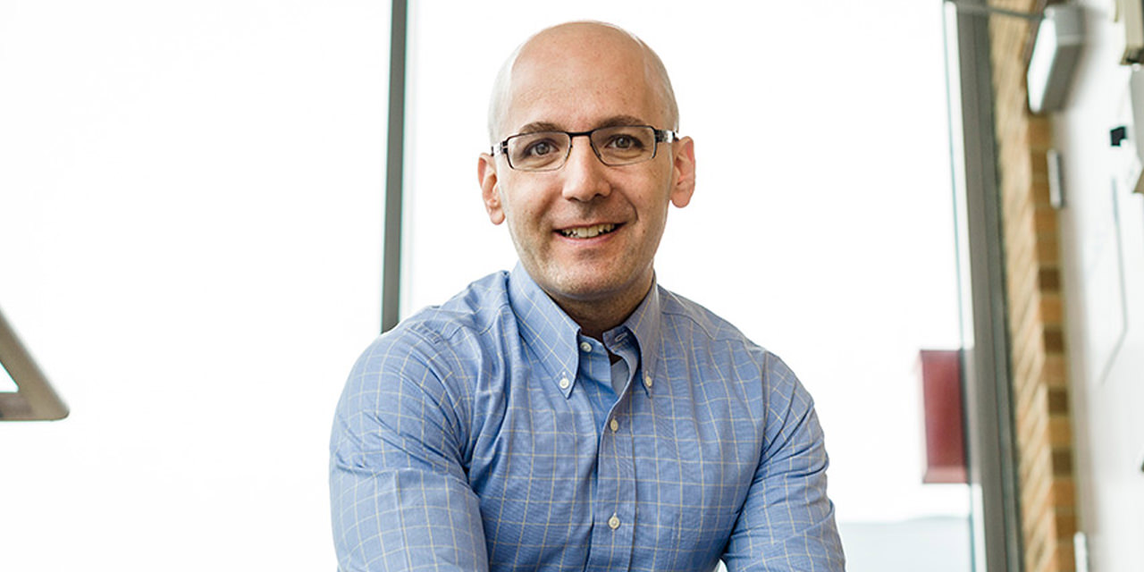 Benjamin Goldstein, sitting down and smiling. He's wearing a blue button-up and glasses.