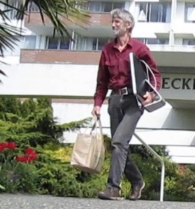 Bruce in a red shirt, walking outside with his computer under his arm.