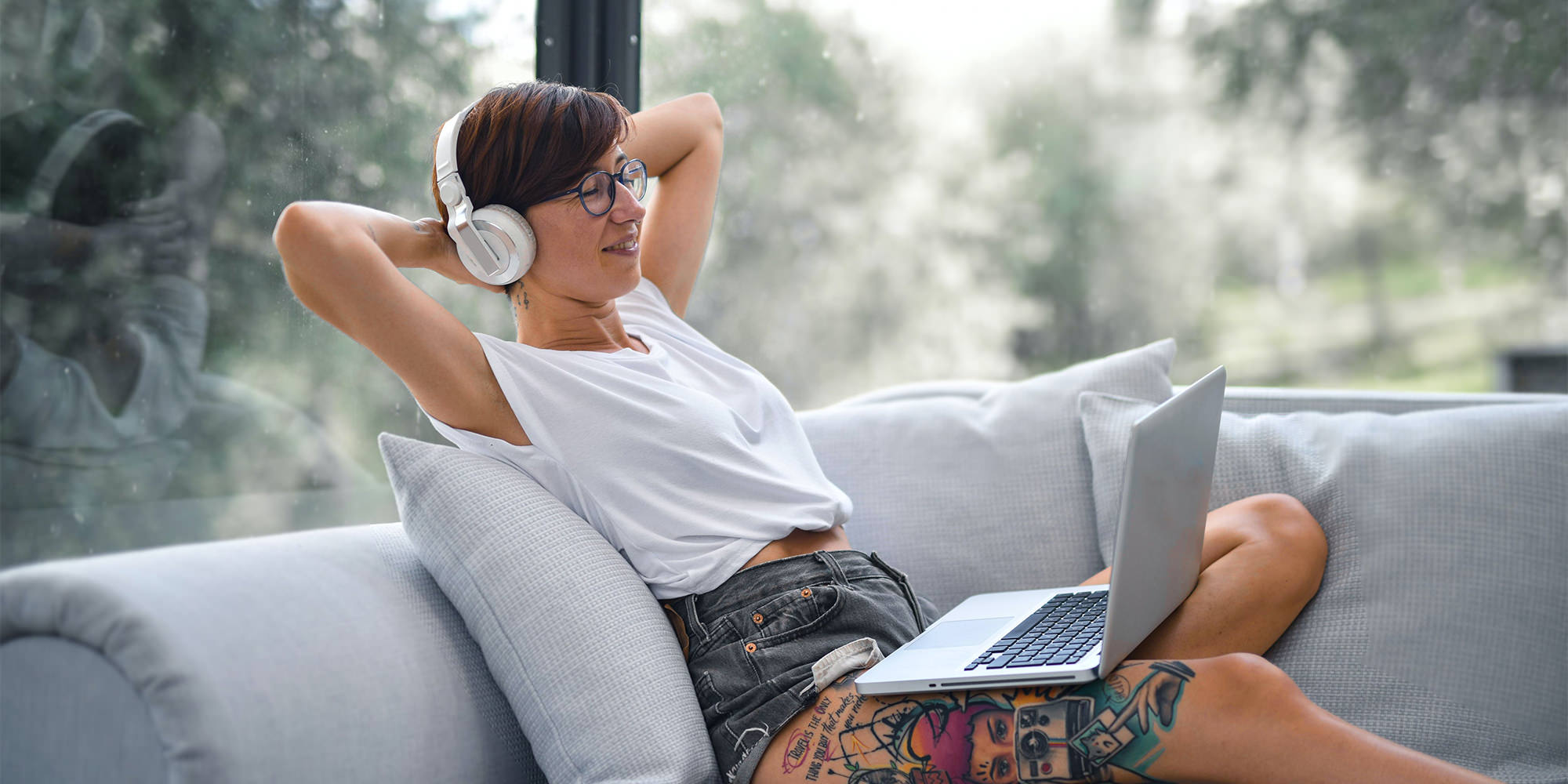 A woman in her late twenties with short hair and glasses, leaning back and listening to music on a white couch. Her eyes are closed and she looks relaxed - it's music self-care!