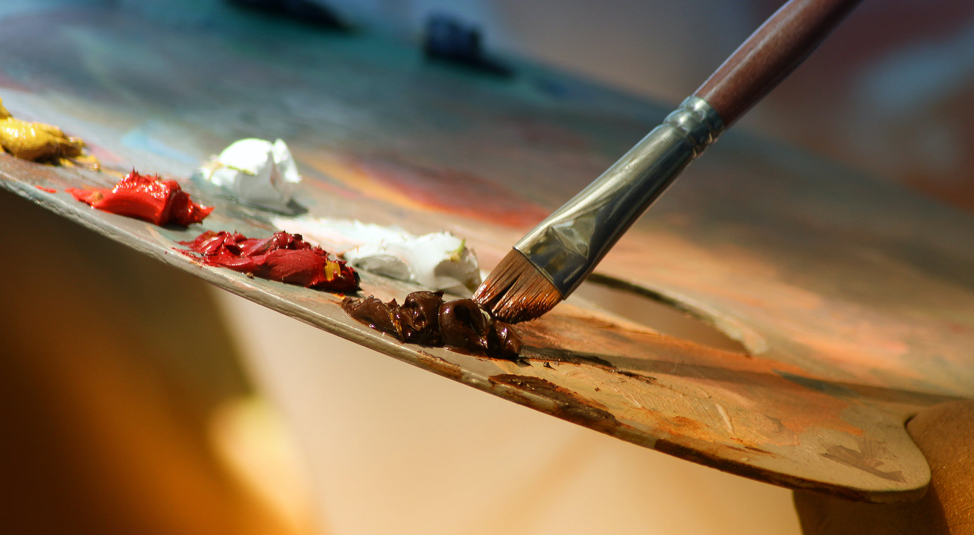 A close-up shot of a wooden paint mixing palette, with a brushing dipping into brown paint.