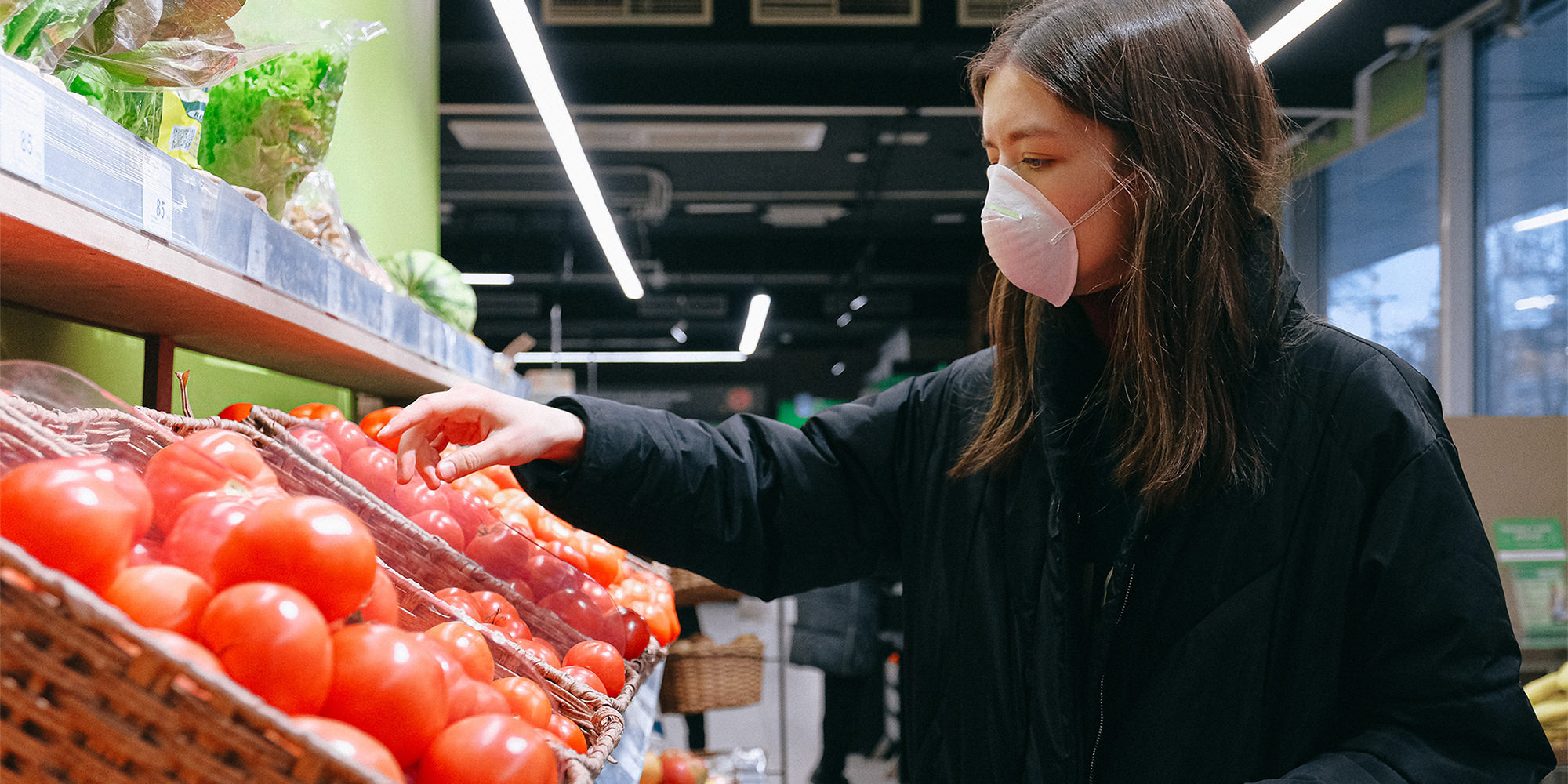 A woman in at the grocery store wearing a face mask. She's reaching towards a basket of tomatoes.