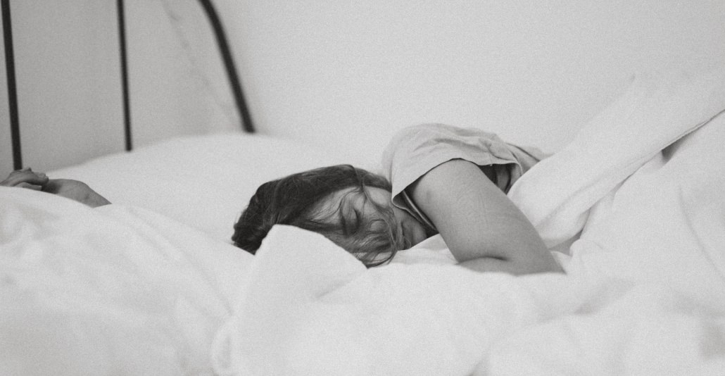 A black-and-white picture of a woman laying in bed, eyes closed and covers bunched up around her.