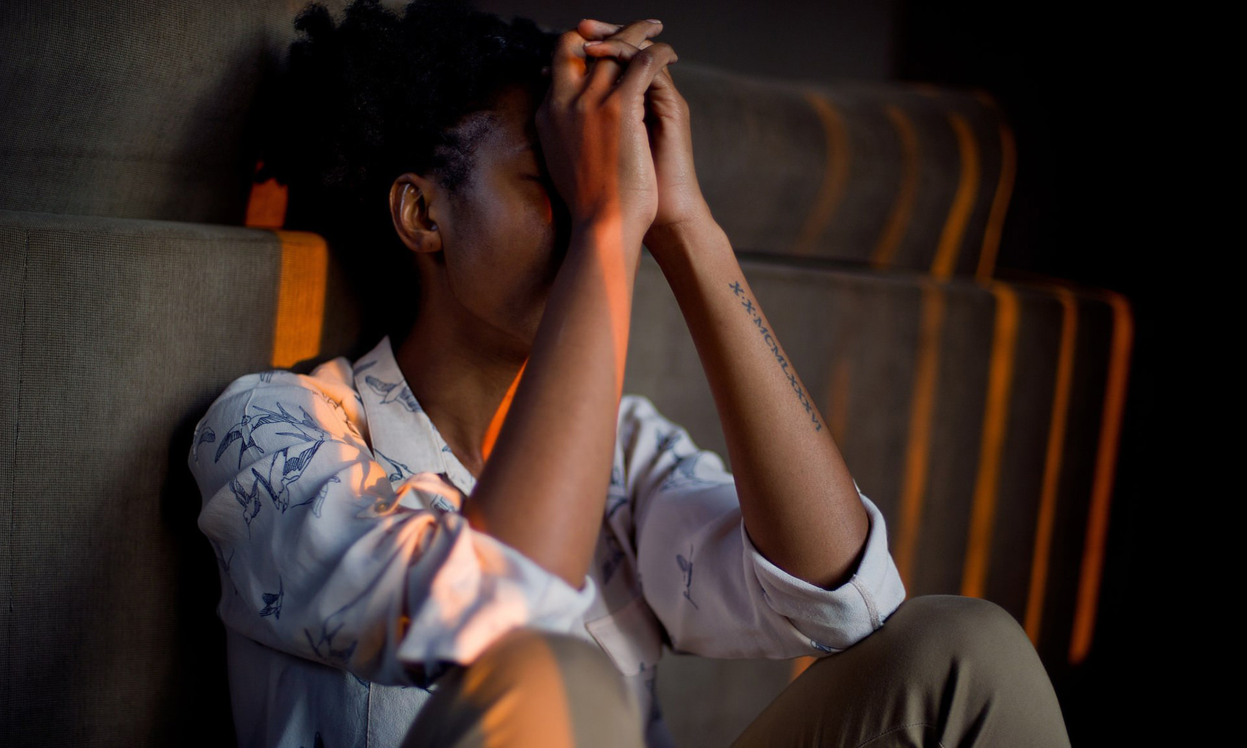 A young African American man sits on the floor, leaning against a couch. He is pressing his clasps hands to his face and looks as if he is under mental strain.