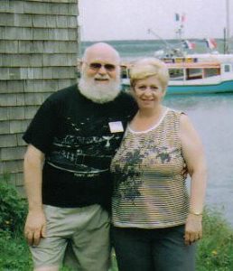 Raymond standing with his wife, Louise. They are outdoor near the water, and a boat can be seen in the background.