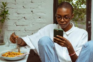 A young black woman with short hair and glasses is sitting at the dinner table with her phone in her left hand. There is a bowl of spaghetti on the table to the left.