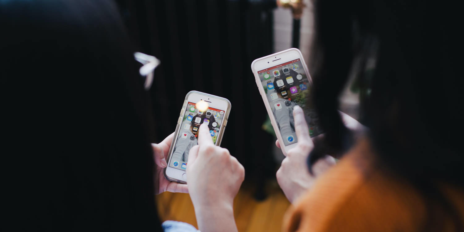 Two young women with long dark hair are shown from the back, holding iPhones.