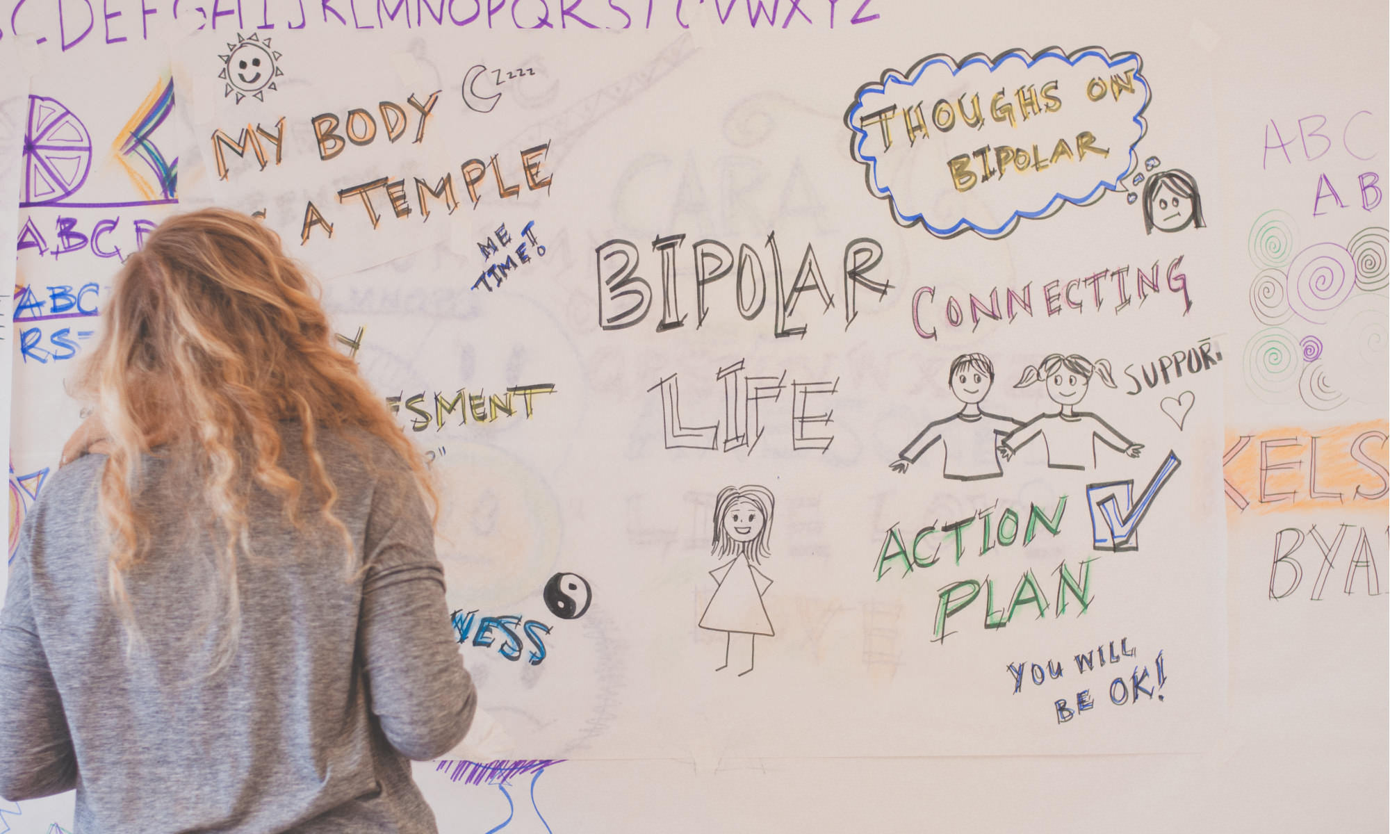A young woman with long blond hair is facing away from the camera towards a huge wall-mounted sheet of paper. She has written and drawn on the paper. There are pictures of people together, a sun, and messages like, "My body is a temple", "Action Plan", and "You will be ok!"