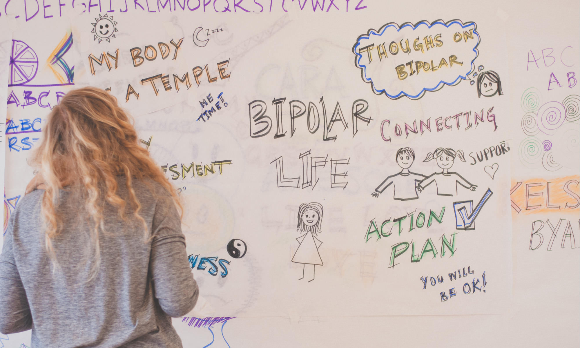 A youth peer researcher on the Bipolar Youth Action Project is facing away from the camera in front of a whiteboard. She is doing graphic facilitation. She has long blonde curls.