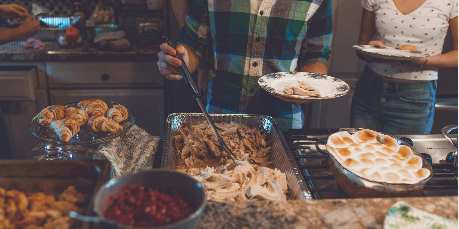 Two young white people are serving themselves Thanksgiving dinner. The lighting is dark and the people's faces aren't shown.