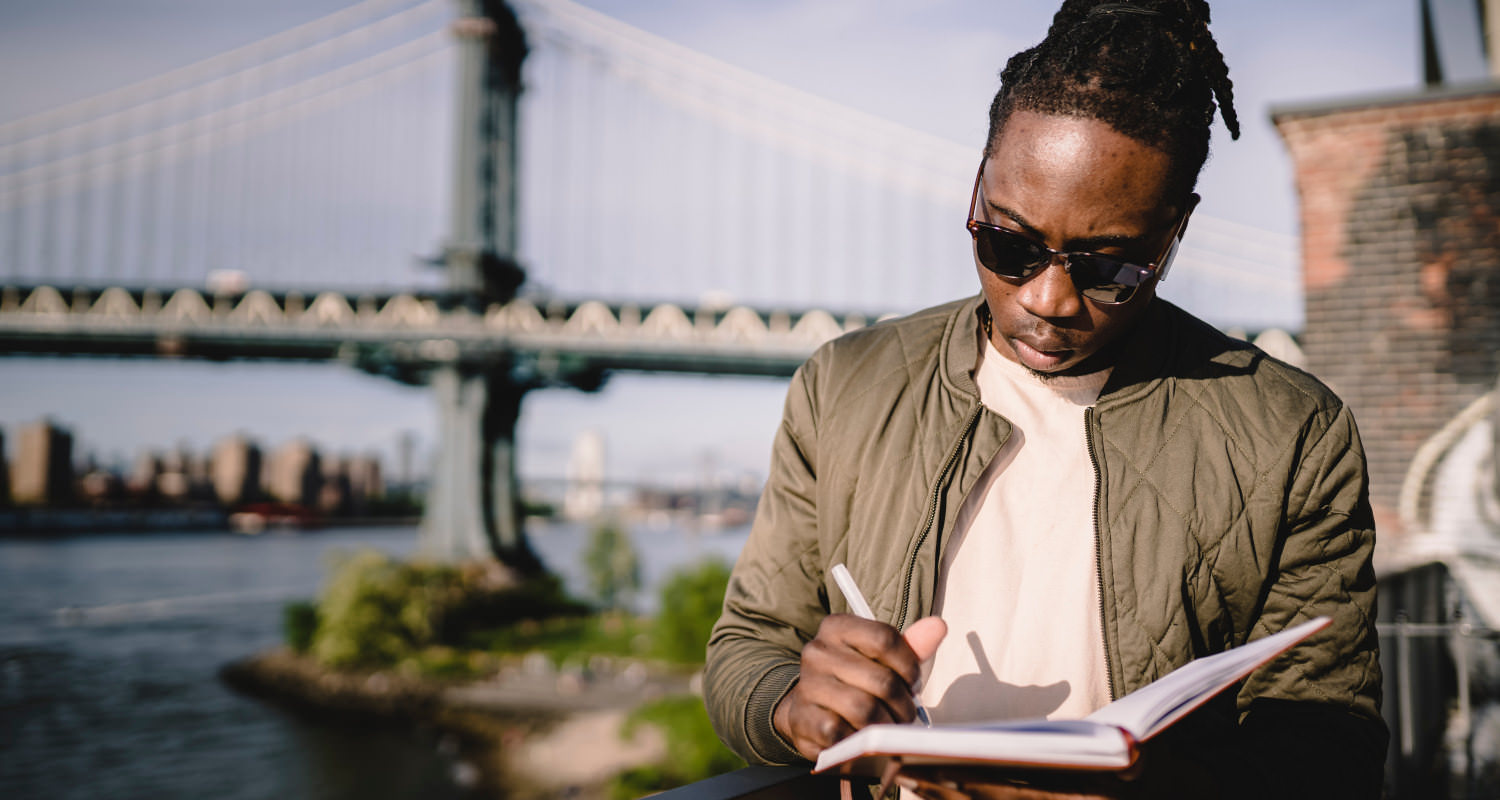 A young black man is holding a notepook and a pen, and is looking down in concentration. He has long, dreadlocked hair that is tied up, is wearing sunglasses, and is standing outdoors near a bridge.