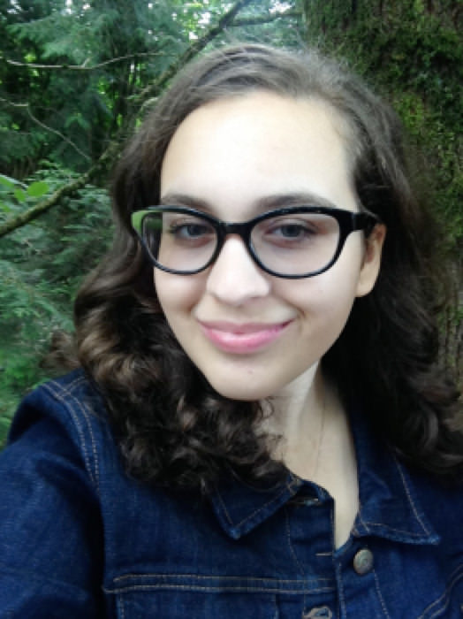 A heatshot of Mikaela. She has long brown curled hair and is wearing a jean jacket and thick-rimmed black glasses with a bit of a vintage look. She is outside in the forest and is turned slightly to the side, smiling.