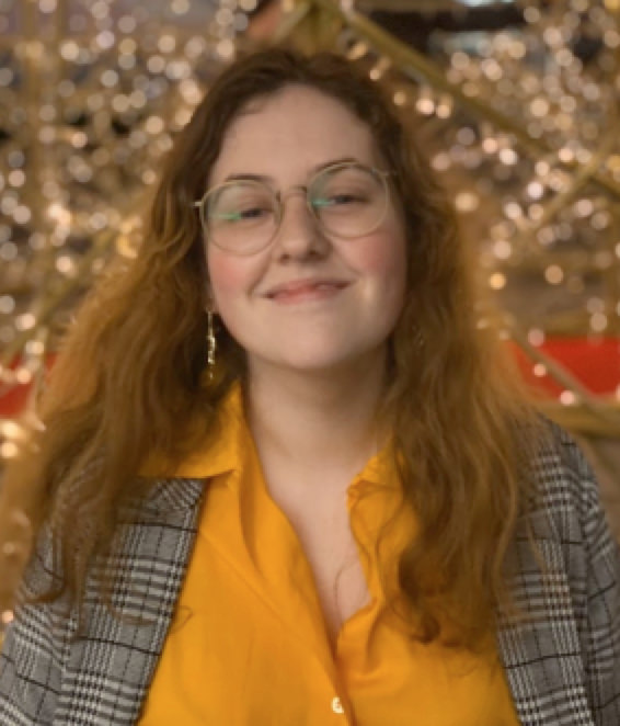 A headshot of Theresa. They have long, reddish brown hair and are wearing a mustard yellow button-up, a plaid overcoat, and round glasses with thin rims. They look to be outside; there appear to be leafless trees with Christmas lights strung on them in the background, blurry and out of focus. They have their head upturned slightly and are smiling.