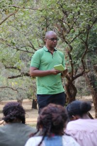 Patrick is standing at an outdoor event in front of an audience. He is wearing a green shirt and sunglasses, and is reading from a small book.