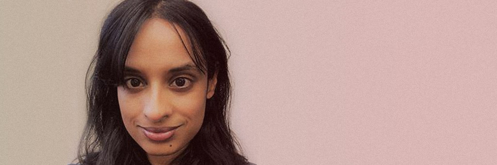 A headshot of Trisha against a pinkish tan background. She is looking slightly downwards and is smiling with a closed mouth. She is South Asian and has long black hair and brown eyes.