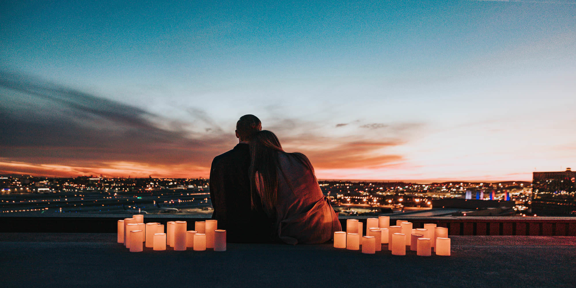 A silhouetted man and woman sit on a building rooftop at sunset. She is resting her head on his shoulder, and they are faced away from the camera, looking out over the city skyline. They are surrounded by large lit candles.