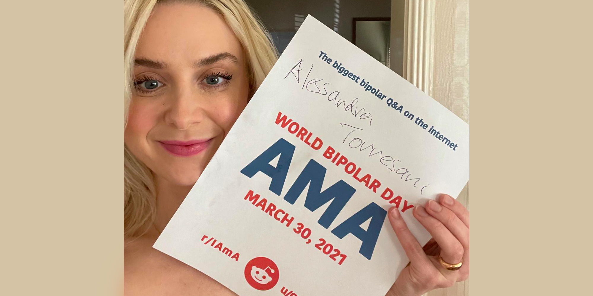 Alessandra indoors. She has white skin, shoulder-length blonde hair, and blue eyes. She is holding the AMA proof sign and smiling with a closed mouth.