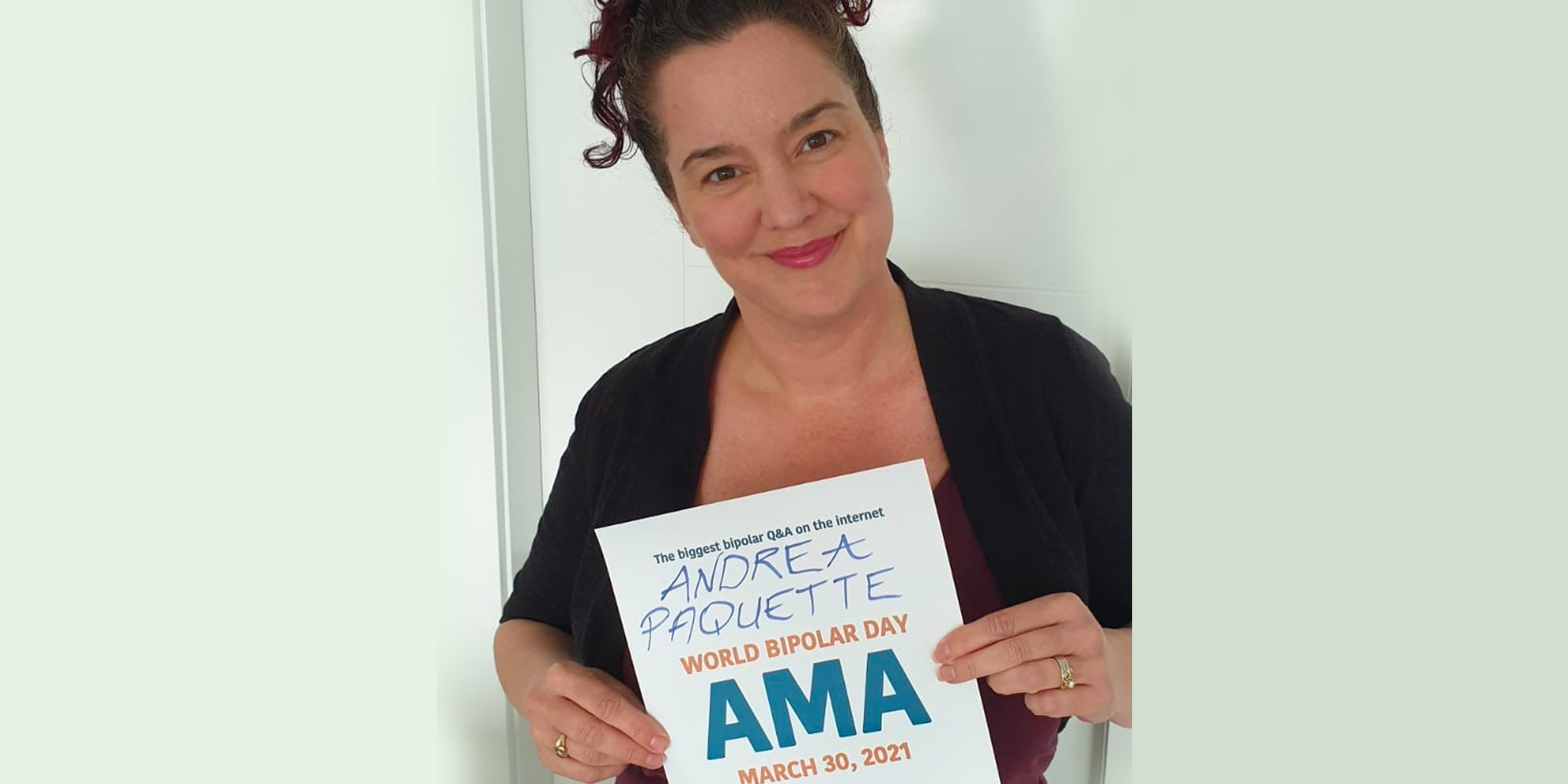 Andrea is wearing a black cardigan and holding an AMA proof sign with her name written on it. She is Caucasian and has dark curly hair with red tips. She is smiling warmly.