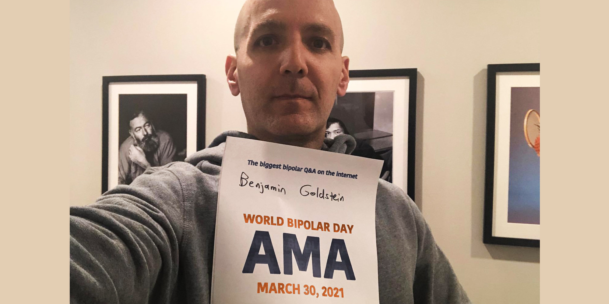 Benjamin Goldstein, is standing indoors, against framed grayscale photographs. He is white and bald and is wearing a grey hoodie. He's holding the AMA proof sign with his name printed on it.