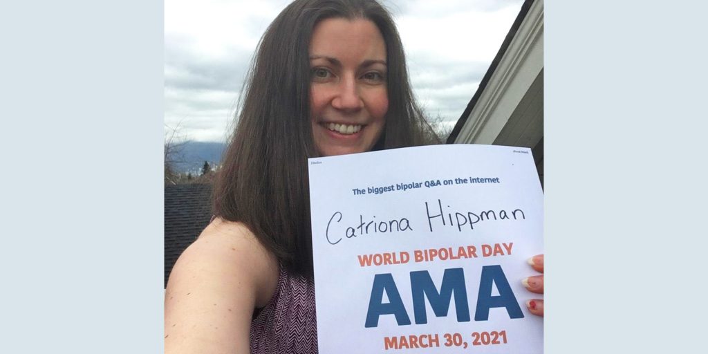 Catriona is standing outdoors holding a proof sign with her name on it and smiling. She is Caucasian and has brown hair and eyes, and is wearing a short-sleeved pink shirt.