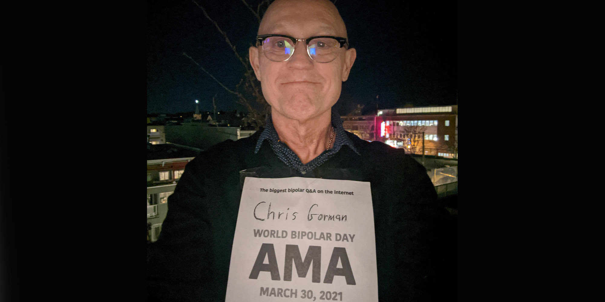 Chris is standing up outdoors at night, perhaps a higher-floor balcony. Buildings with lit windows are in the background. He is Caucasian and is bald and wearing horn-rimmed glasses and a dark sweater with a navy blue collar showing from his shirt underneath. He is holding the AMA proof sign.