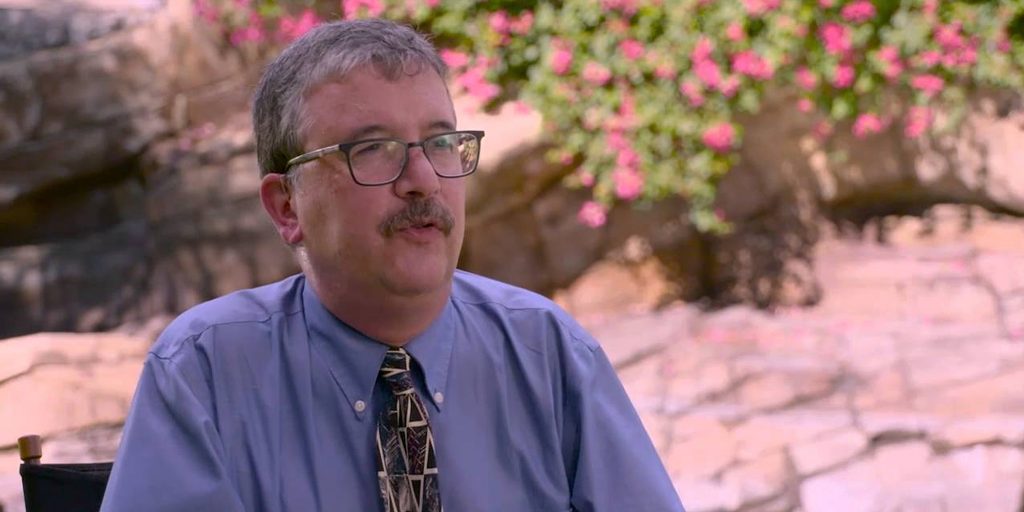 David is sitting outside in front of a sandy-coloured natural wall with a large bush hanging over, with bright pink flowers. He is Caucasian has dark grey hair and a moustache. He is wearing dark-rimmed rectangular glasses, a light blue dress shirt, and a brown tie with arrow patterns.