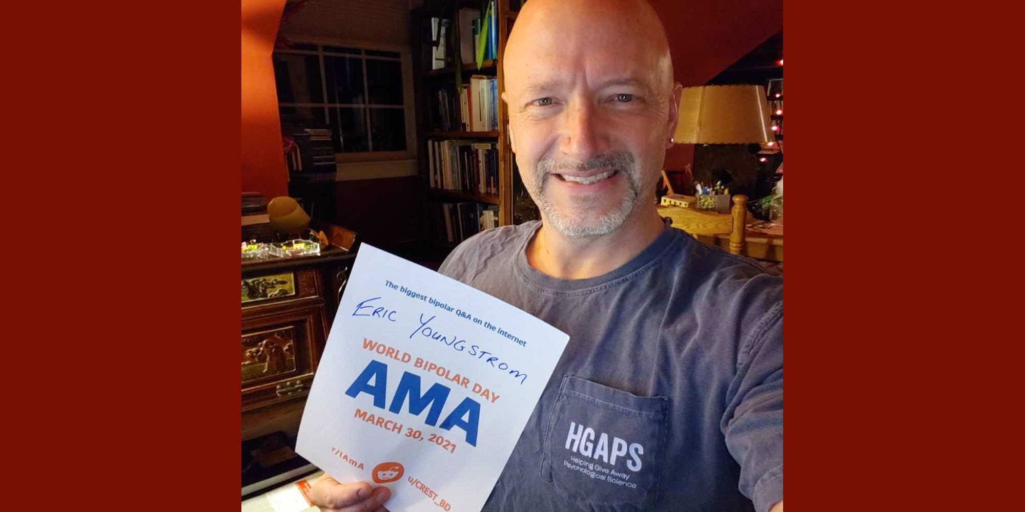 Eric is in a room in his home that looks like it might be an office, with reddish and white walls, dark-stained furniture, and a bookshelf. He is Caucasian and bald with a short greyish beard, and is wearing a grey shirt with HGAPS written on the pocket. He is holding the proof sign.