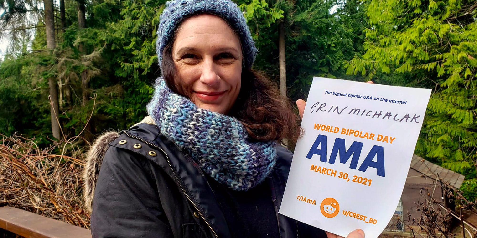 Erin outdoors in a forest. She is Caucasian and has curly dark brown hair. She's wearing a blue knitted hat with a matching scarf and a black jacket. She is smiling warmly and holding a sign proving she will be involved in the bipolar ama, with her name and the AMA date and time on it.