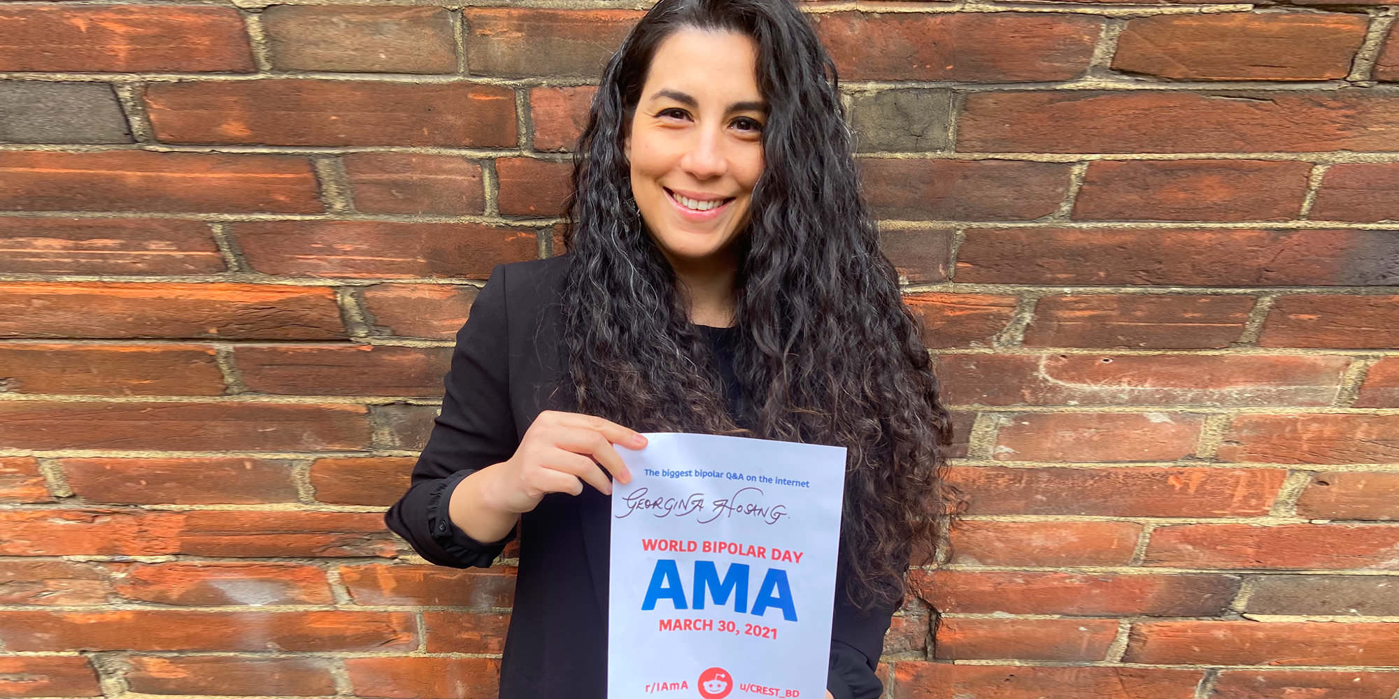 Georgina is standing outdoors in front of a brick wall and is holding the proof sign. She has tan skin and long, curly black hair. She is wearing a dark shirt or blazer and smiling.