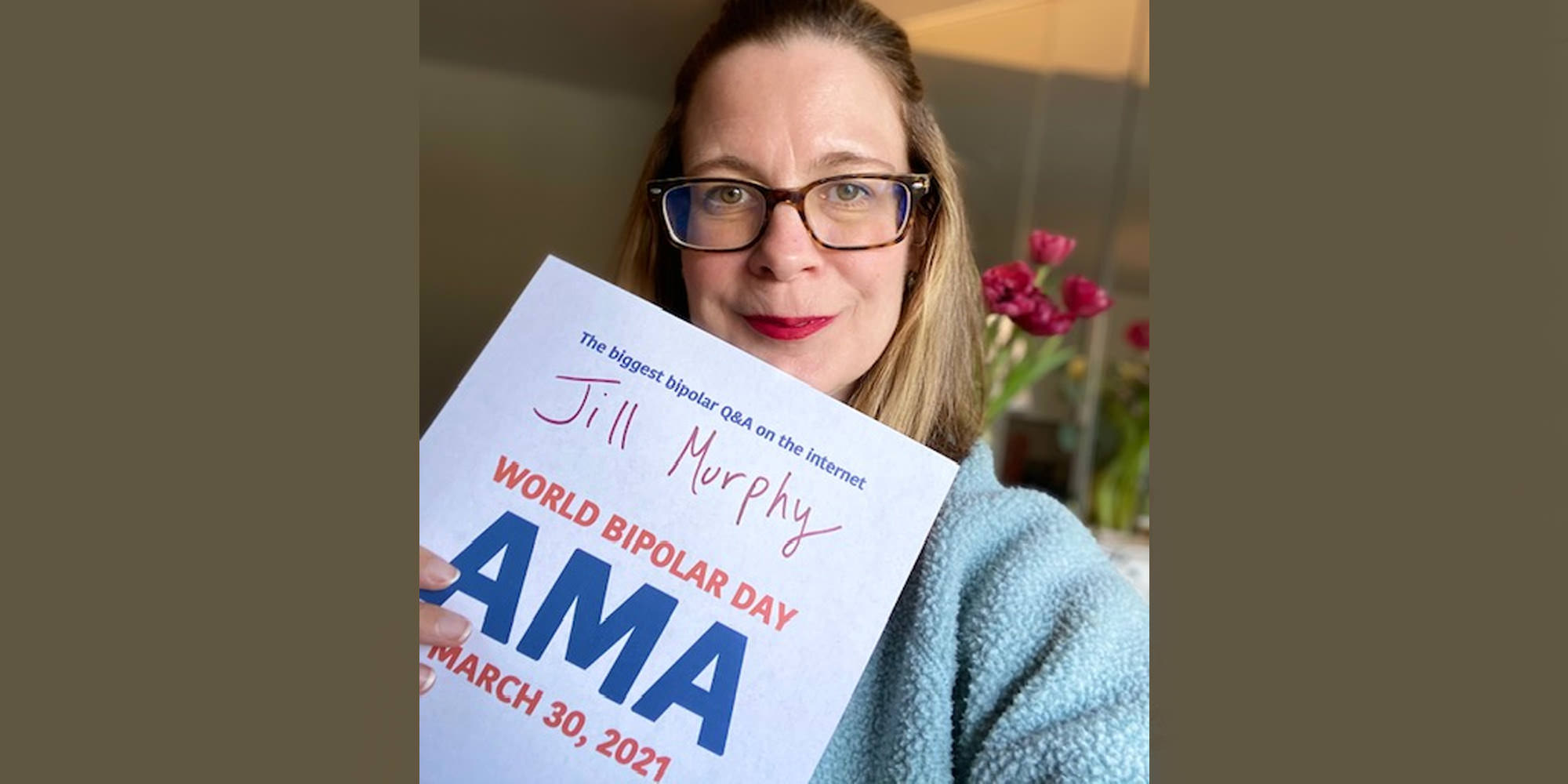 Jill is standing in a room with a large mirror and red tulips. She is Caucasian, has shoulder-length ombred hair, and is wearing thick-rimmed tortoiseshell glasses and a fuzzy light blue sweater. She is holding the AMA proof sign.