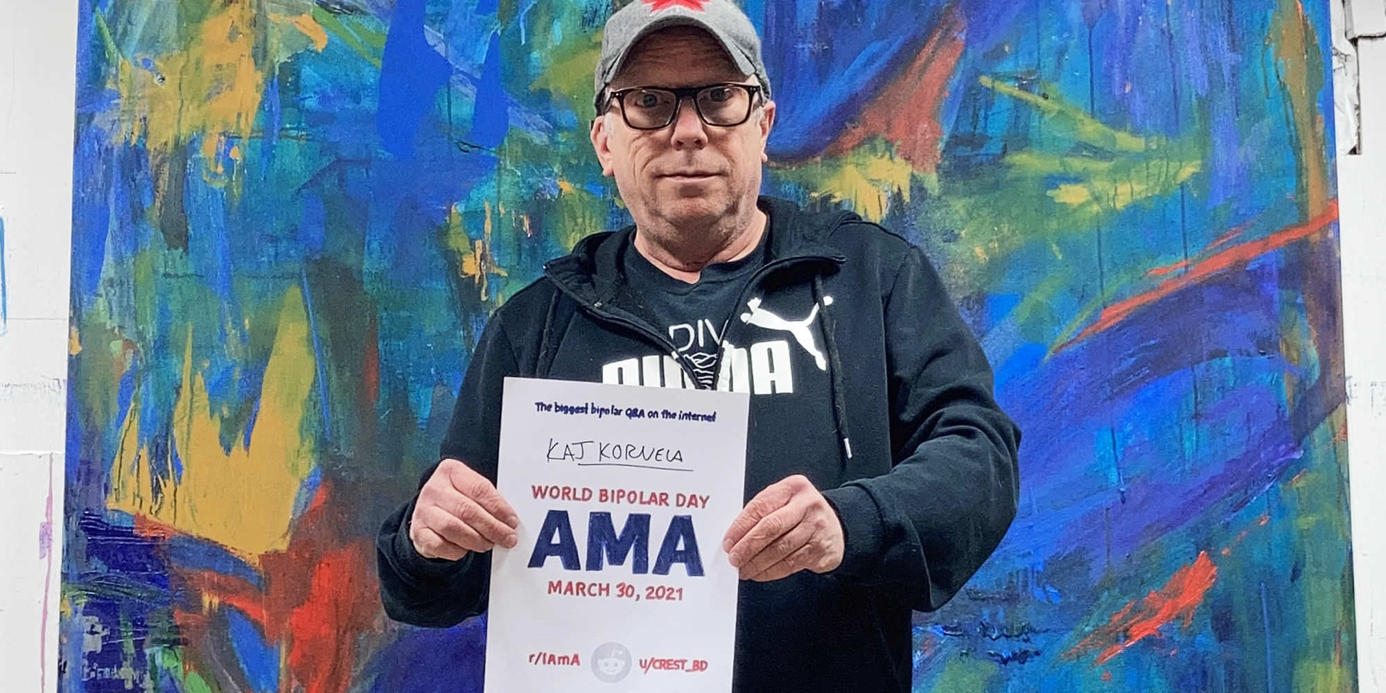 Kaj is standing in front of a huge, abstract painting of his. It's largely blue with big splashes of red and yellow. He is Caucasian and is wearing thick-rimmed glasses, a grey baseball cap, and a black Puma hoodie over a Joy Division t-shirt. He is holding a proof sign with his name.