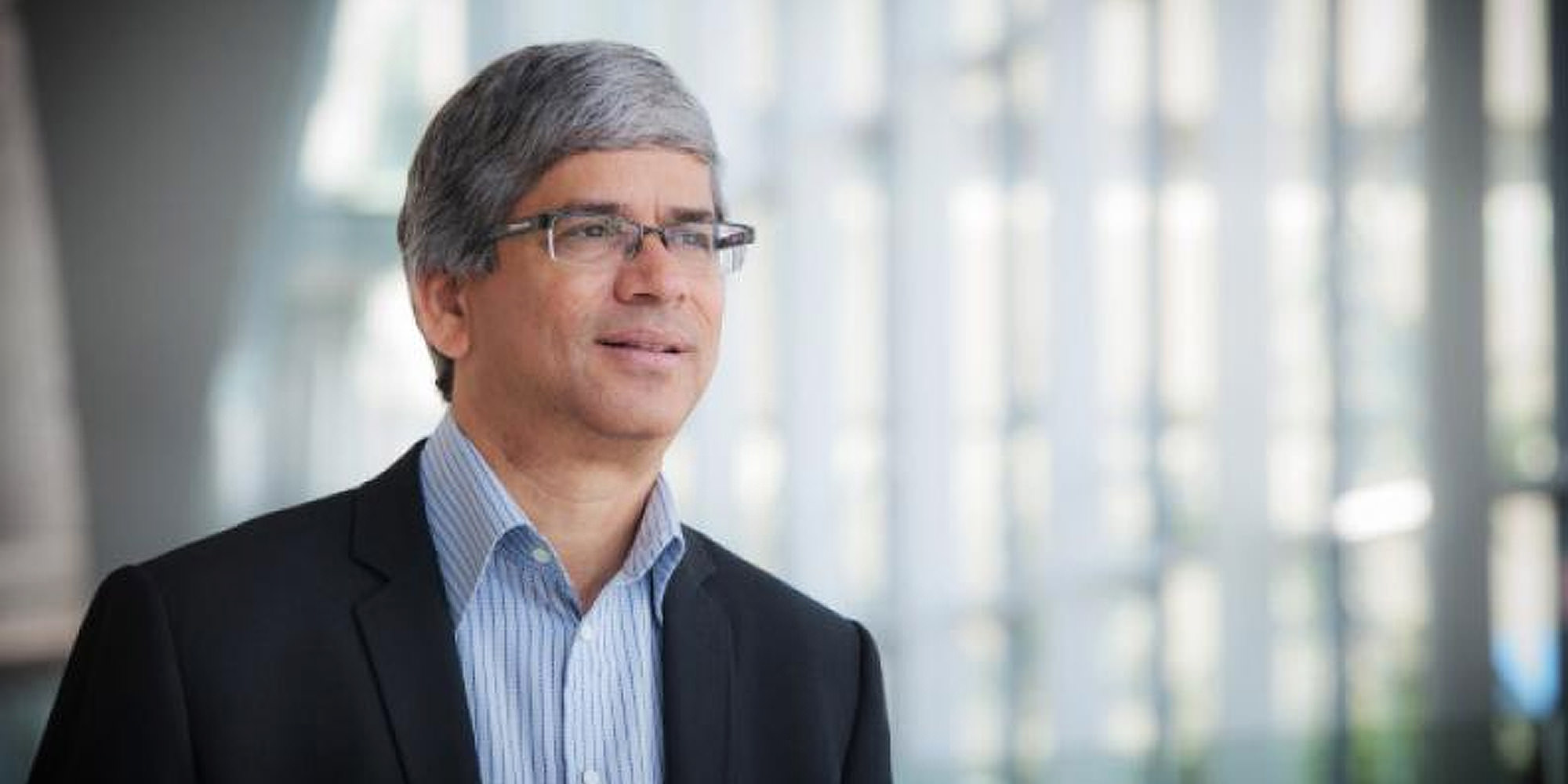 Lakshmi is standing indoors in a well-lit room. He is South Asian and has grey hair and rectangular glasses. He is wearing a striped blue-and-white shirt and a navy blue suit jacket and has a thoughtful expression on his face.