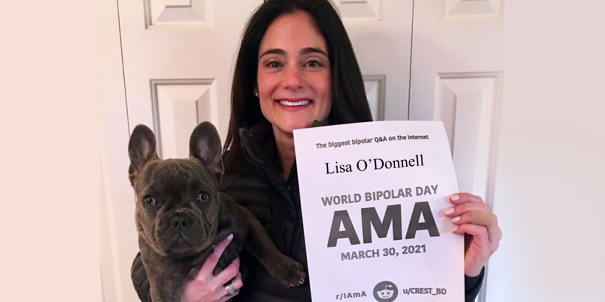 Lisa is standing indoors in front of a closet door, holding her dog! Her dog is a dark grey frenchie. Lisa is Caucasian and has long black hair, she she is holding a proof sign printed in black and white. 