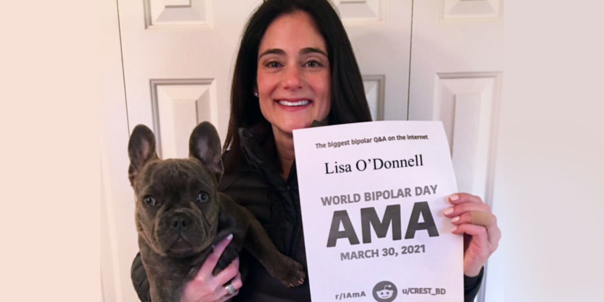 Lisa is standing indoors in front of a closet door, holding her dog! Her dog is a dark grey frenchie. Lisa is Caucasian and has long black hair, she she is holding a proof sign printed in black and white.