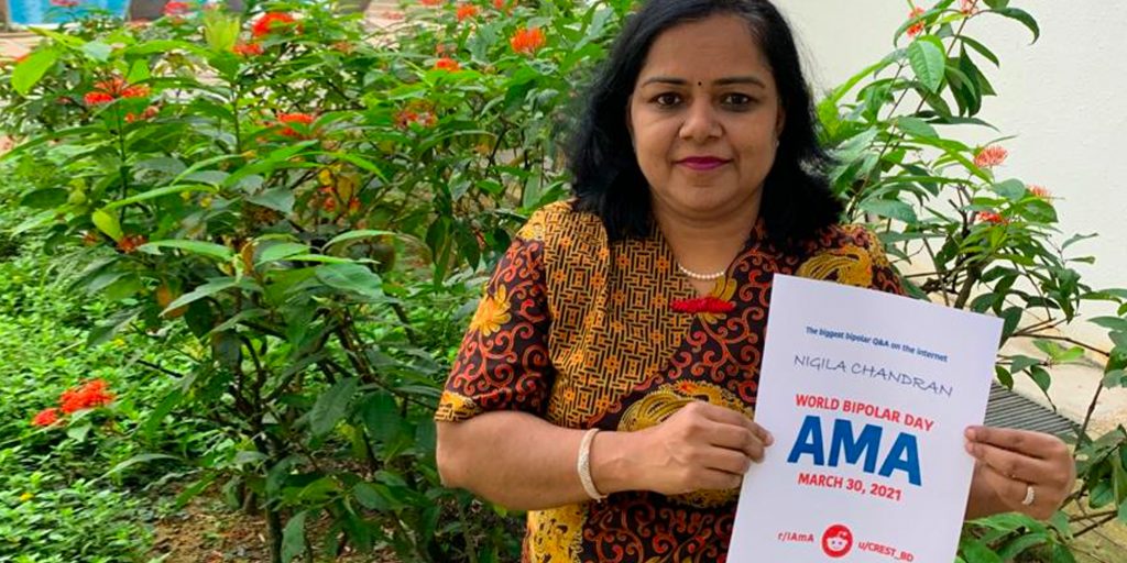 Nigila is standing outdoors in front of a tropical-looking bush with bright red berries. She is South Asian and has shoulder-length dark hair. She is holding an AMA proof sign with her name written on it, and is wearing a yellow and brown patterned dress with red accents.