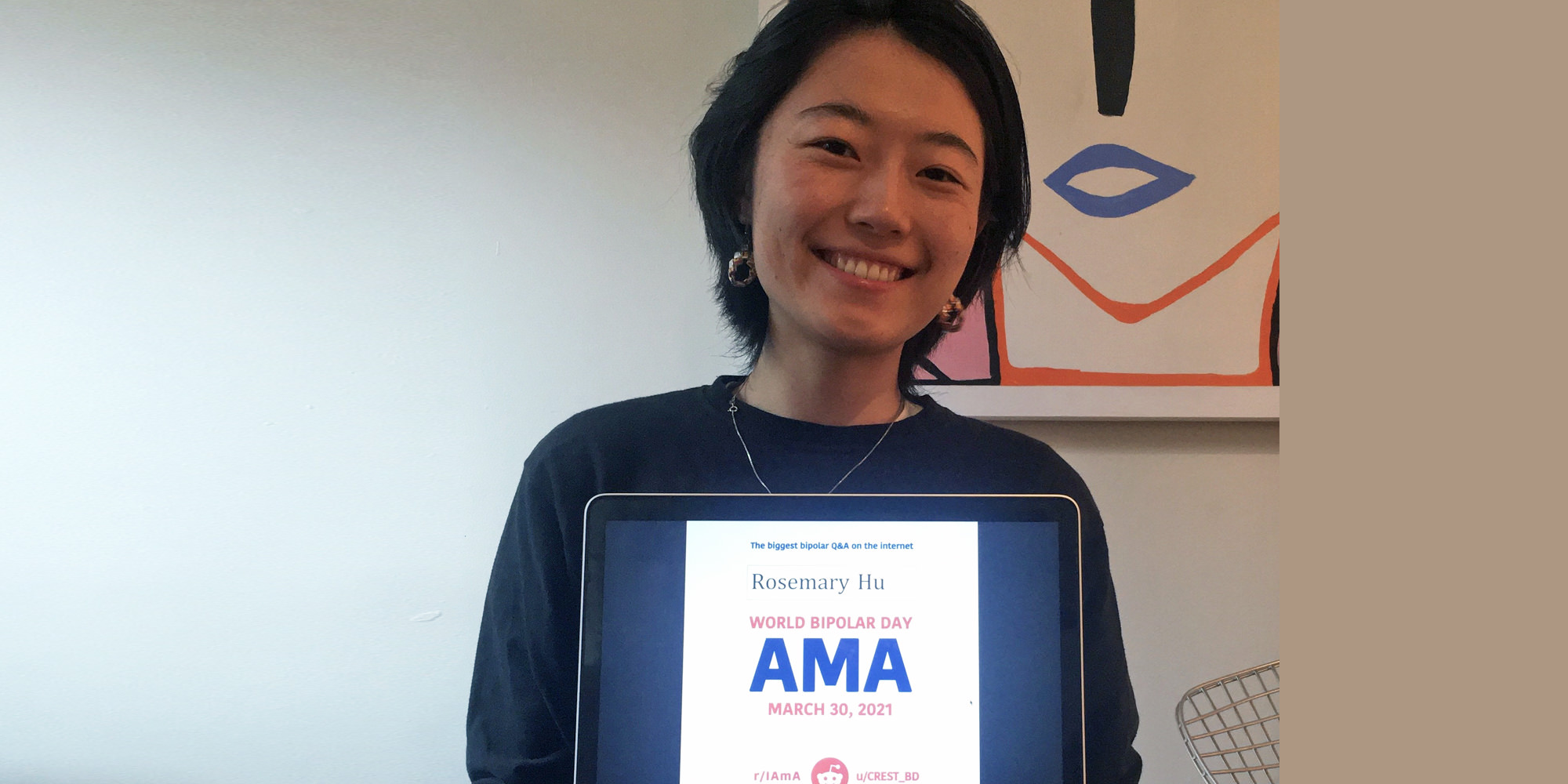 Rosemary is standing in front of a minimalist, modern painting of a woman's face. She is Chinese Canadian and is holding an iPad or laptop screen with the AMA proof and her typed name up on the screen. She has short black hair, gold earrings, and is wearing a lightweight dark sweater.