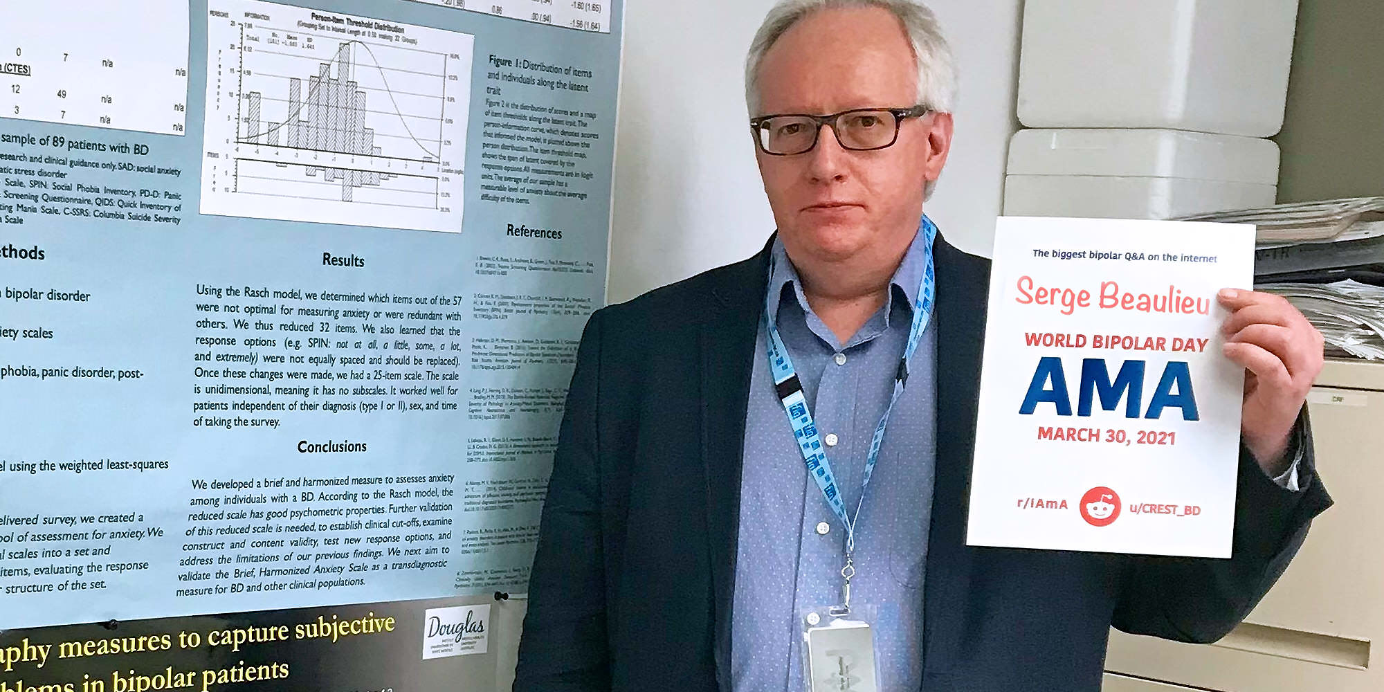 Serge is standing next to a study poster and looking into the camera with a neutral expression. He is Caucasian and has grey hair and thin-rimmed oval glasses. He is wearing a grey suit with a blue shirt and a lanyard. He is holding the AMA proof sign.