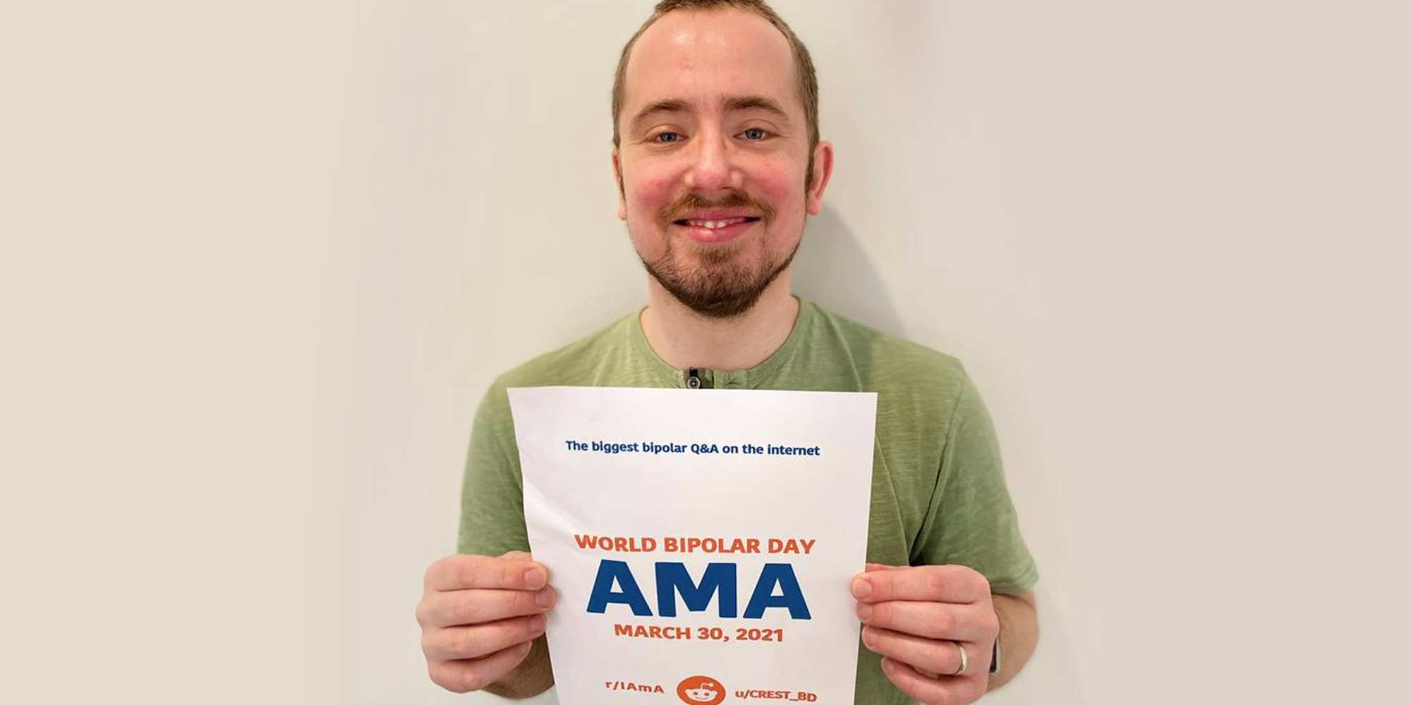 Thomas is standing in front of a beige walling holding up the AMA proof sign. He is Caucasian, with short, close-cropped brown hair and a slight beard. He is wearing a green t-shirt.