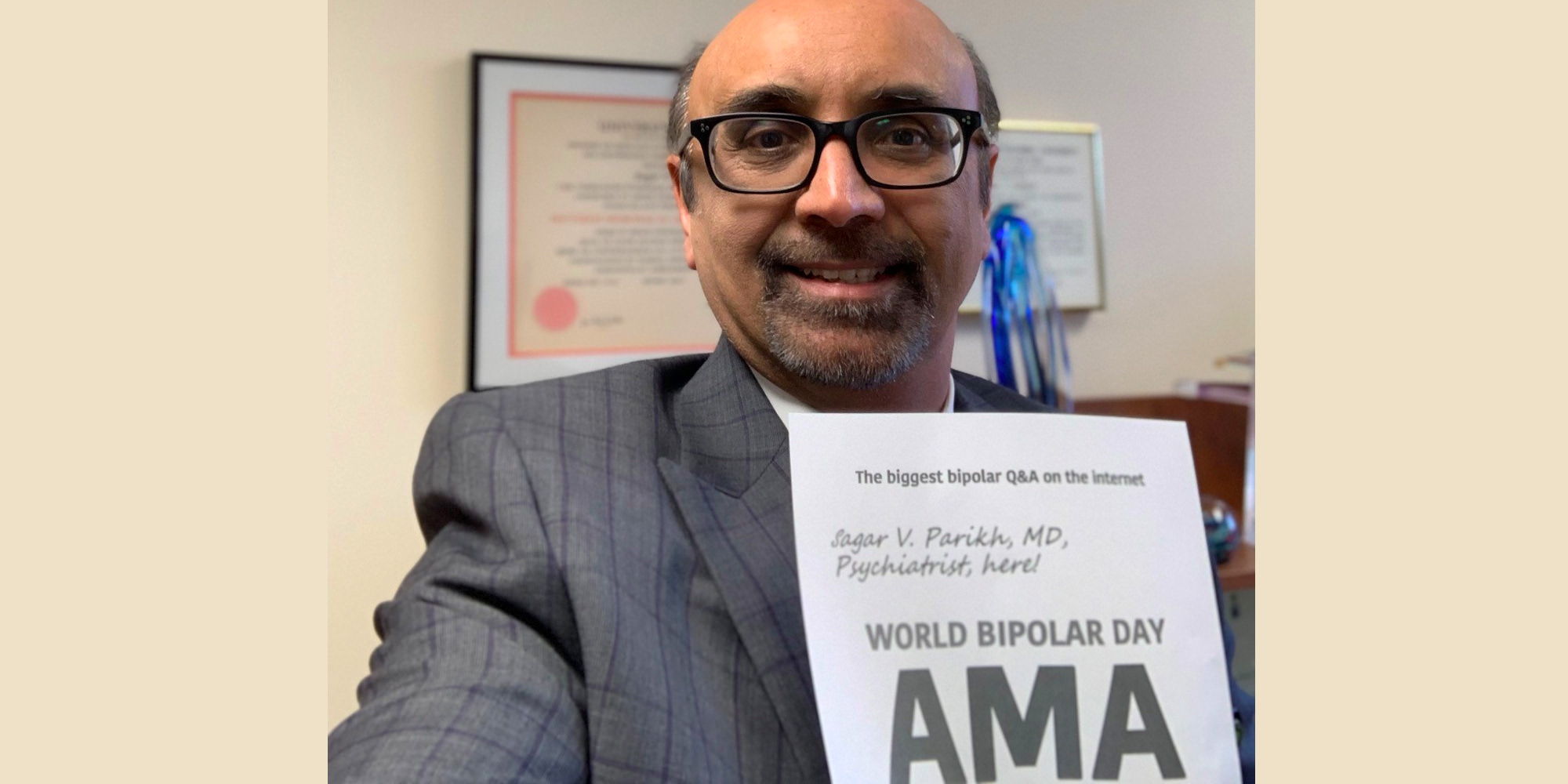 Sagar is in his office holding the AMA proof sign with his name written on it. He is Southeast Asian and is wearing a grey suit with a grid pattern and black, rectangular, thick-rimmed glasses.