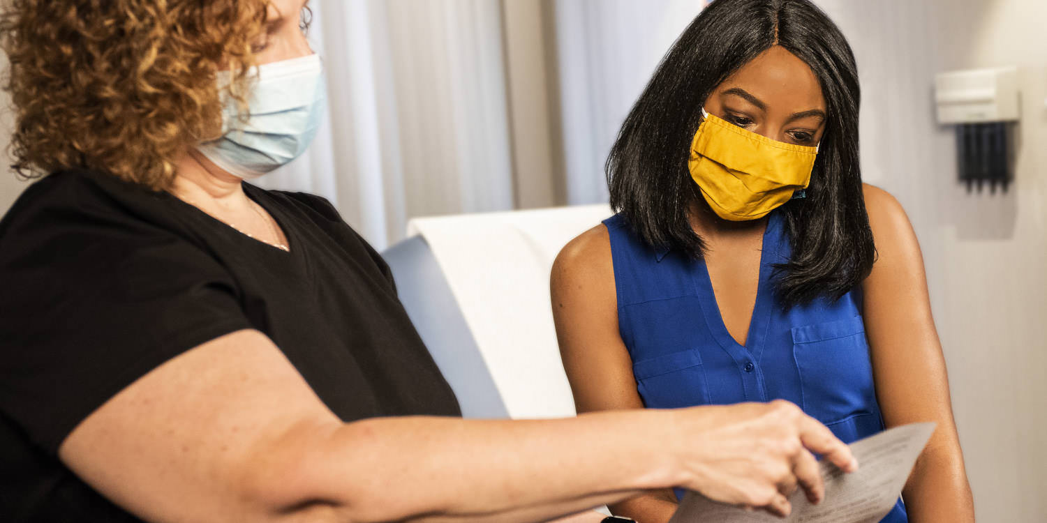 Two women in a health-care setting. The one on the left, a Caucasian woman with curly red-brown hair and a standard issue medical mask, is showing a paper of some sort to a darker-skinned woman with shoulder-length black hair wearing a yellow mask.