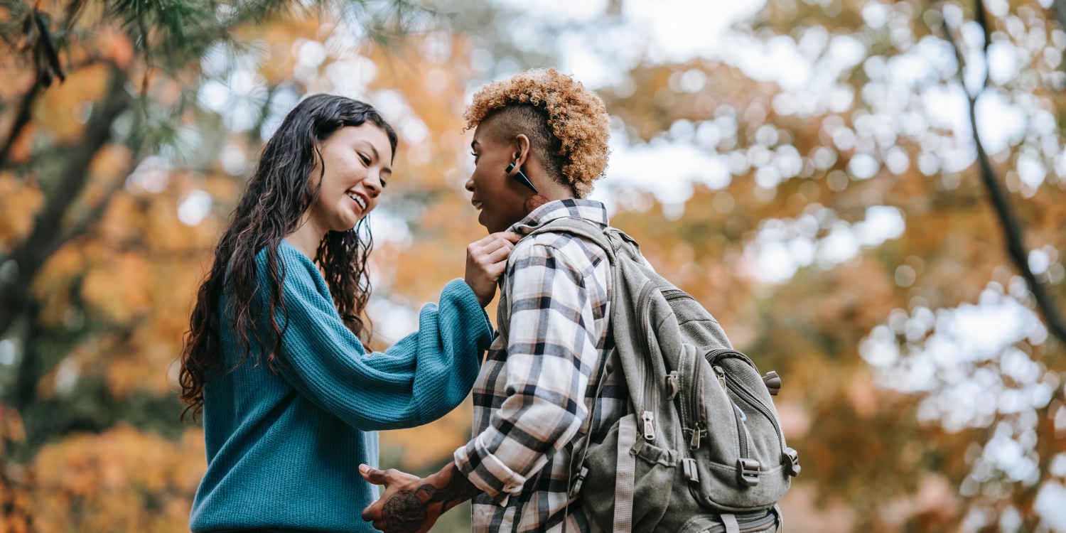 Two young women together in autumn. On the right is a young black woman with orange dyed hair with the side cropped short. She is wearing a black checked jacket and has tattoos and is smiling and appears to be saying something. She is facing her partner, a young South Asian woman with long black hair. The partner is wearing a green sweater and adjusting the black woman's collar. She has an expression of affection on her face.