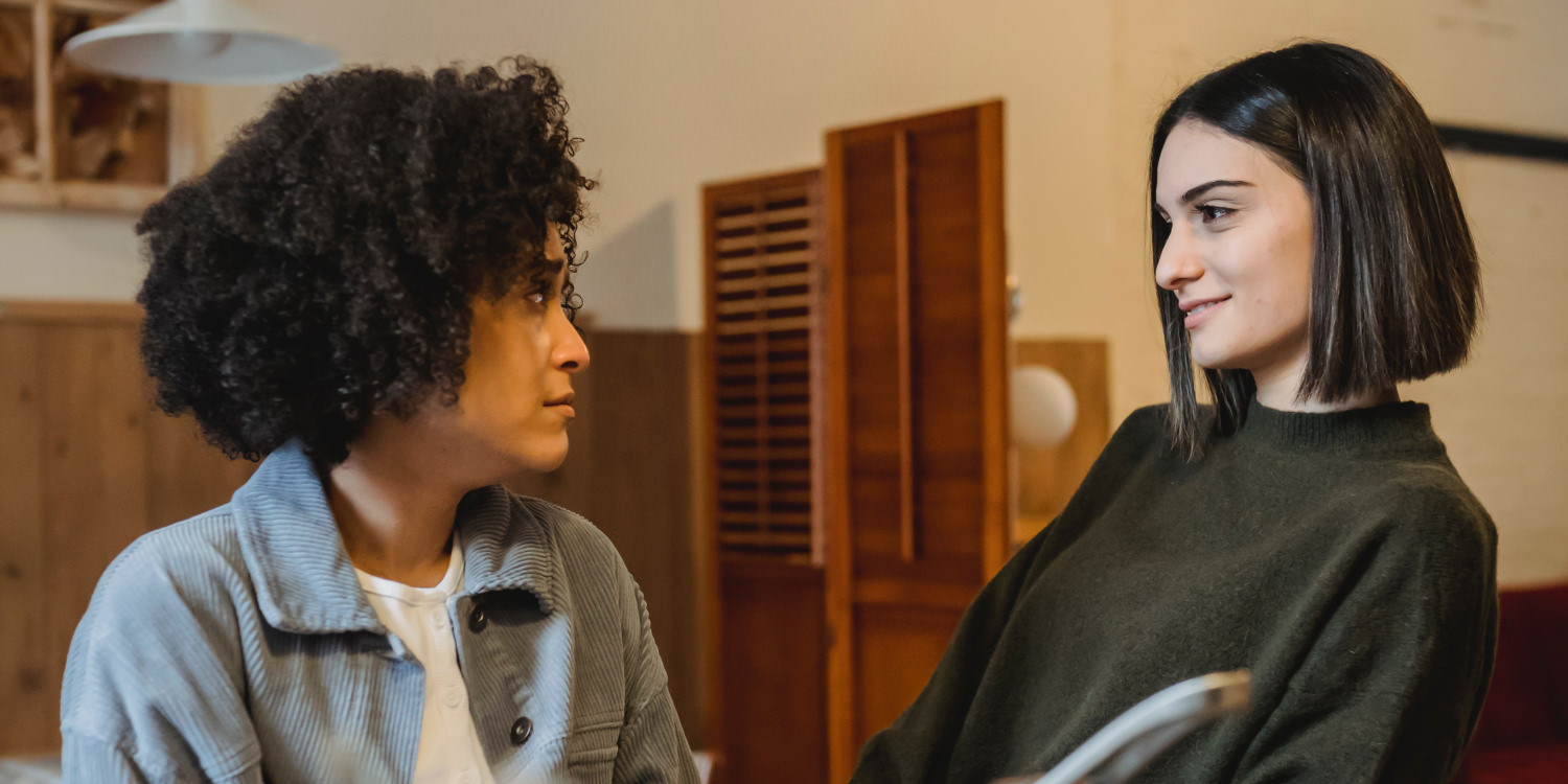 A young black woman is looking at her friend with a worried expression. She has curly black hair and is wearing a blue jacket. Her friend is possibly Latinx or Caucasian and is smiling encouragingly. She has a straight brown bob and a brown sweater on.