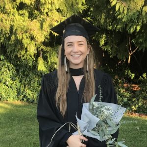 A picture of Nathalie at graduation. She is Caucasian and has long light-coloured hair. She is smiling and holding a bouquet of elegant green and white flowers. Her graduation cap and gown are black and she is wearing two long dangling earrings.