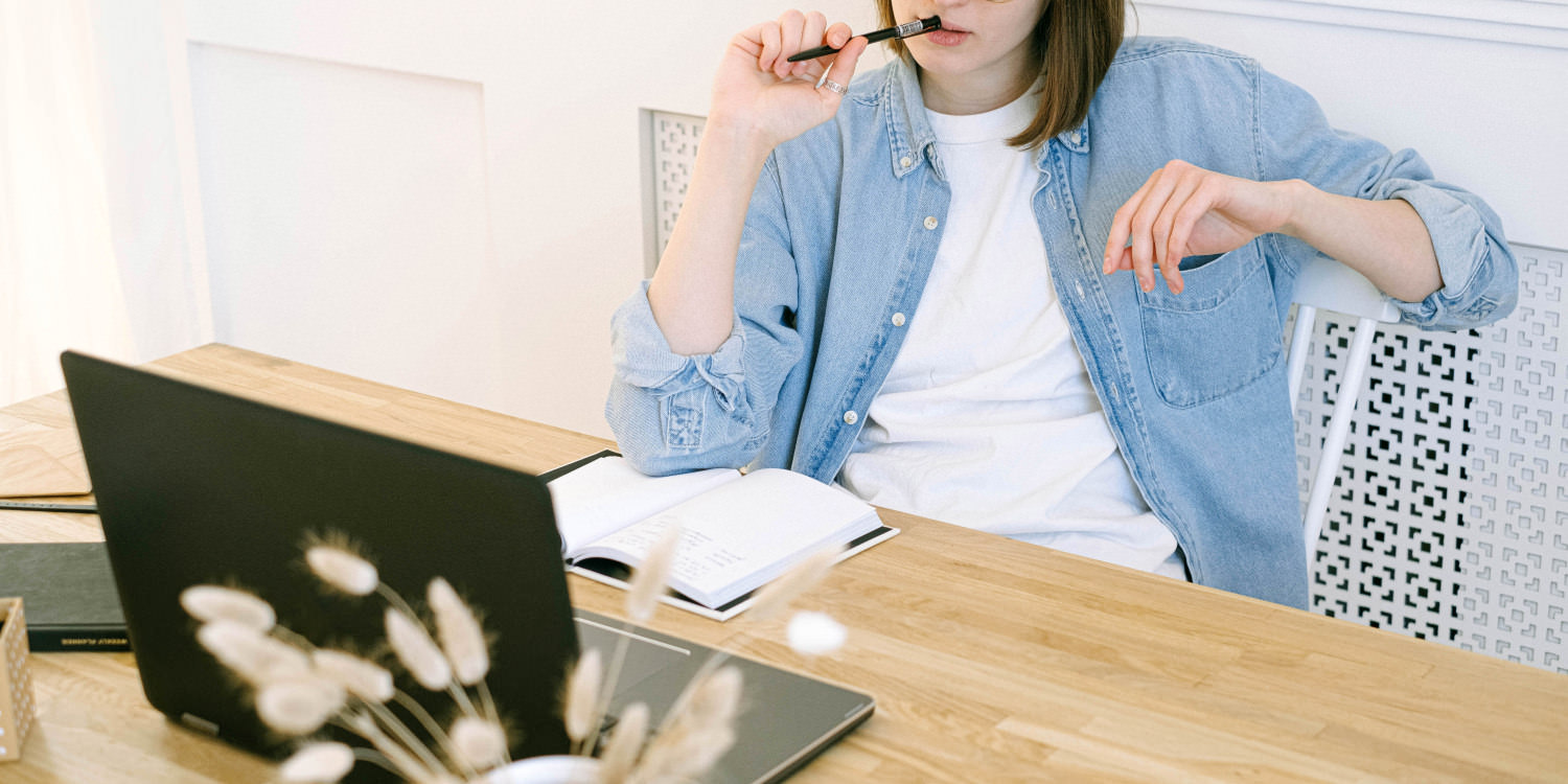 A young Caucasian women is sitting in front of a laptop and notebook and is pressing her pen to her lips in thought. She has shoulder-length brown hair and is wearing a thin oversized jacket that resembles denim. The upper half of her face is not shown in the picture.